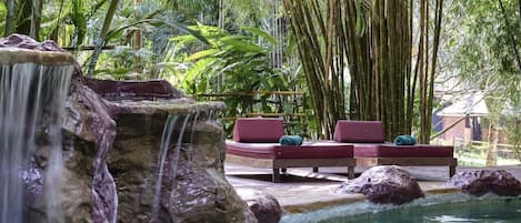 Zona de piscina con cascada, rodeada de vegetación exuberante y sillones de relax rojos.