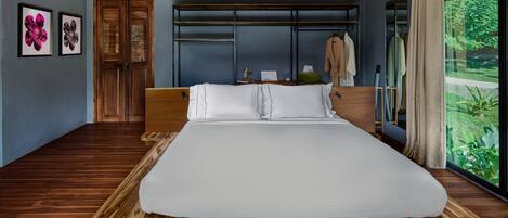 A bedroom with a large bed, wooden flooring, a wardrobe, and a view of greenery through a window.