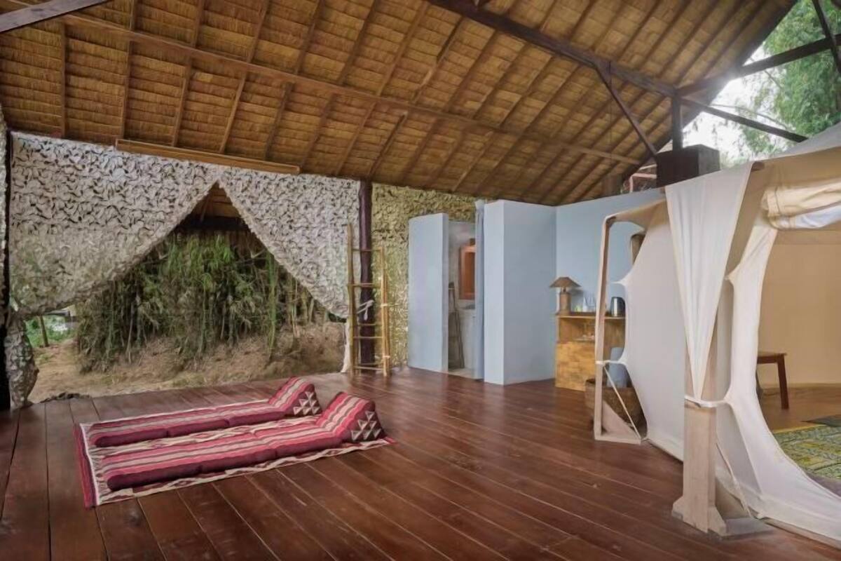 Une pièce avec un plancher en bois, un toit de chaume, un lit avec une literie à rayures et une vue sur la verdure à l’extérieur.