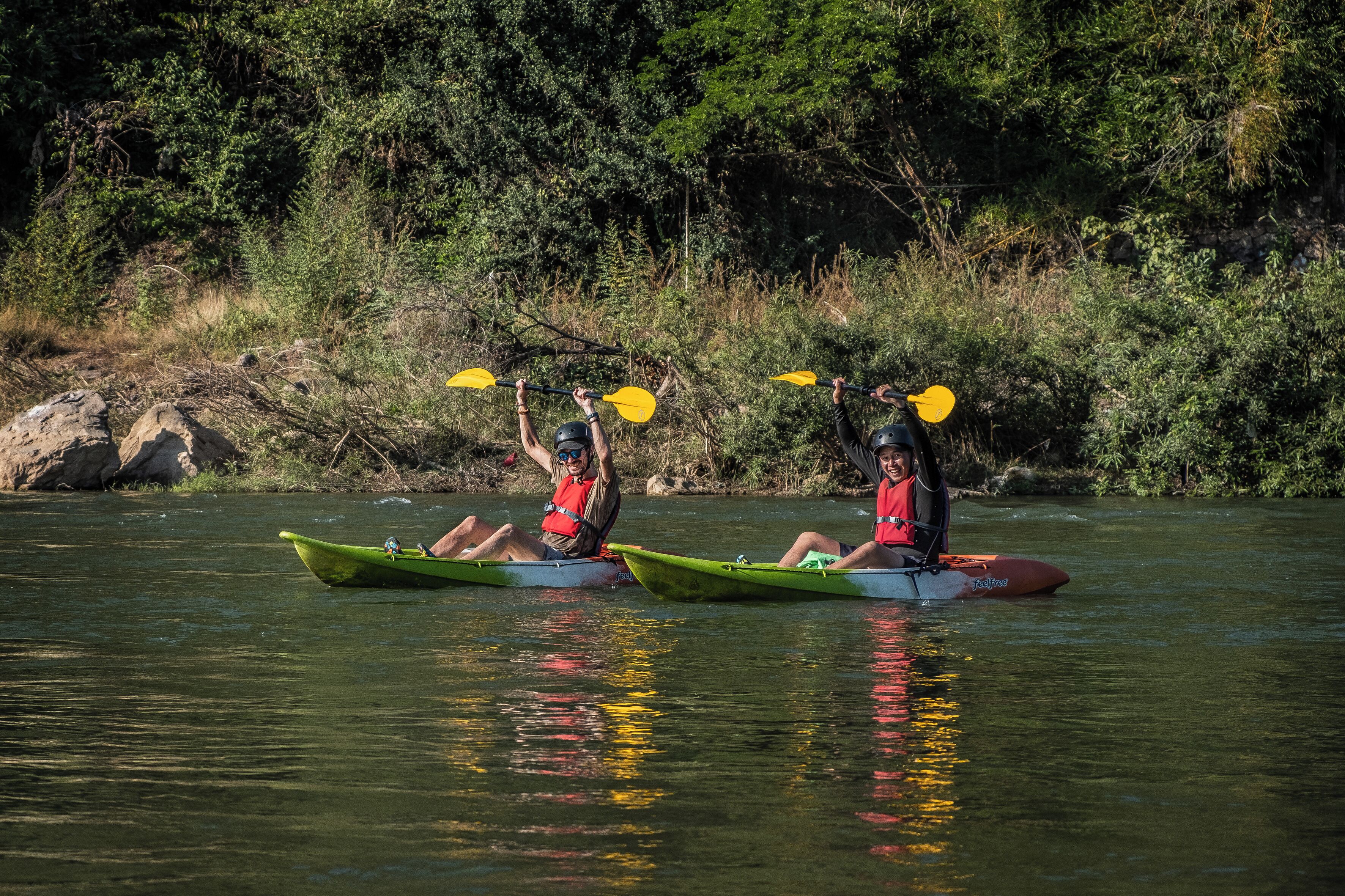 Dos personas remando en kayak por un río tranquilo, rodeado de abundante vegetación.