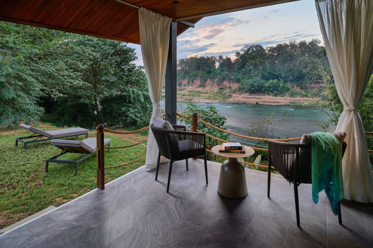 Une terrasse avec vue sur une rivière, entourée de verdure. On y trouve des chaises longues et une table avec un livre.