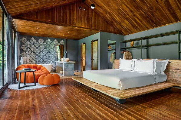 Un dormitorio moderno con techo de madera, una cama grande, sillones orejeros naranja y vistas al exterior.