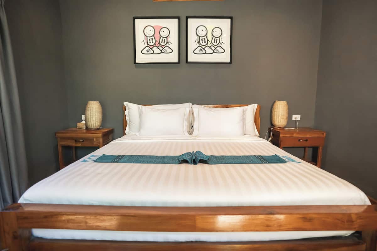 Un lit bien fait, recouvert d’une couvre-lit à rayures et agrémenté de deux oreillers, flanqué de deux tables de chevet en bois sur lesquelles sont posés des vases décoratifs.