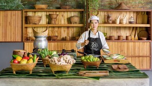 A chef in a kitchen with wooden shelves, baskets of fruits and vegetables, and cooking utensils. - The Namkhan, a Small Luxury Hotel of the World (Luang Prabang)