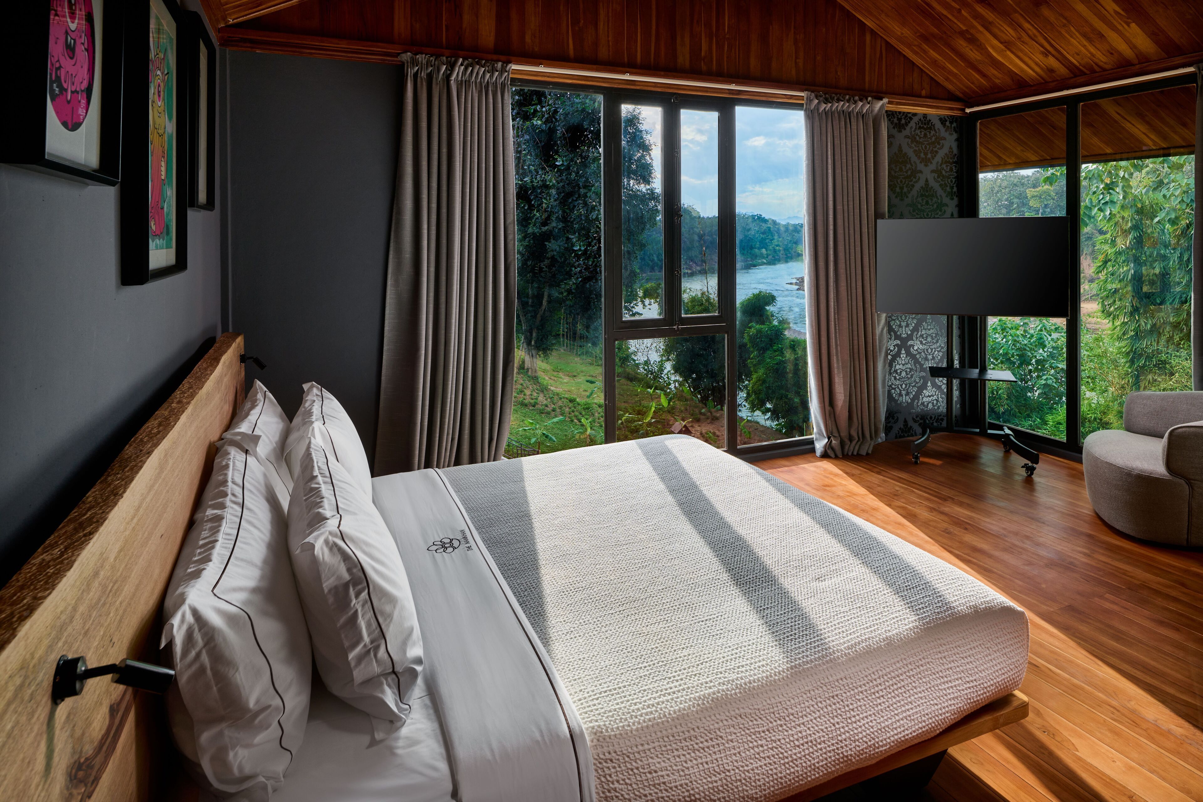 A bedroom with a large bed, a chair, and a TV. It has a view of a river and trees.