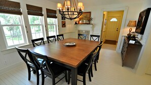 Une salle à manger avec une table et des chaises en bois, de grandes fenêtres équipées de stores, un lustre et une cheminée.