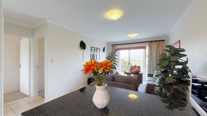 A modern living room with a dark granite countertop, a white vase with orange flowers, a grey sofa, and a large green plant. - Manatoka Coastal Condo (Cape Town)