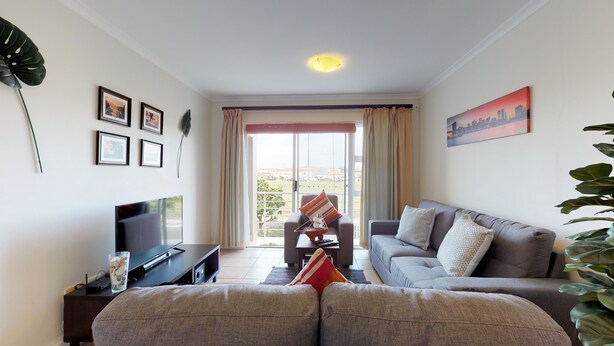 A living room with a sofa, a TV, and a large window with curtains. - Manatoka Coastal Condo (Cape Town)