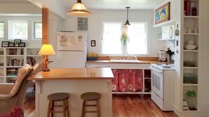 A kitchen with a central island, bar stools, a sink, and a stove. There are windows, a bookshelf, and a potted plant.