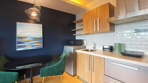Cocina compacta con microondas, tostadora y tetera, armarios de madera, pileta de acero inoxidable y una silla verde.