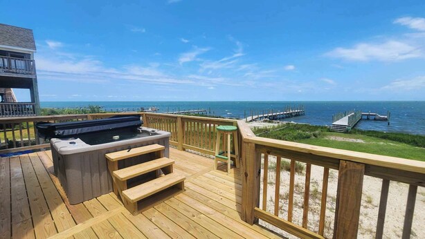 A wooden deck with a hot tub, outdoor seating, and a view of the ocean and pier. - Soundview:  Dog friendly sound front home, den, private dock. (Ocracoke)