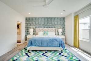 Une chambre avec un papier peint à motifs, une tête de lit capitonnée, un lit recouvert d’une courtepointe bleue, deux tables de chevet avec des lampes et un tapis à motifs géométriques.