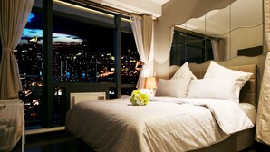 Hotelkamer met een groot raam dat uitzicht biedt op de stad 's nachts. De kamer heeft een opgemaakt bed met witte lakens en een bloemenboeket.