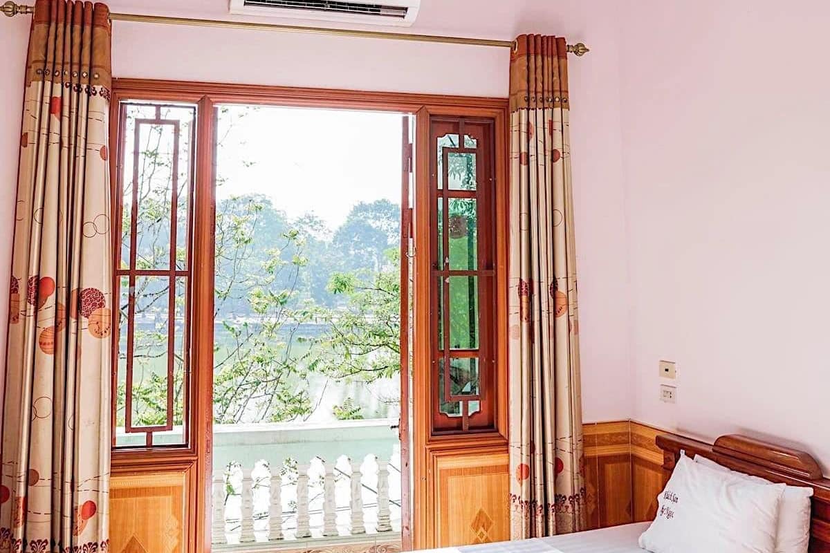 A bedroom with a bed, an air conditioning unit, a window with curtains, and a view of greenery outside.
