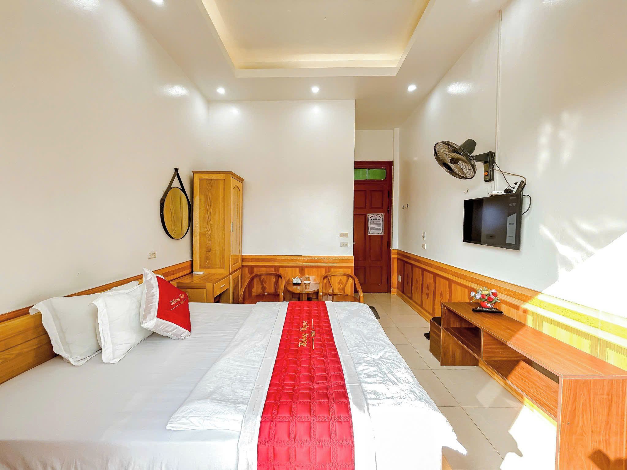 A hotel room with a bed, a wooden wardrobe, a desk with a TV, and a fan.
