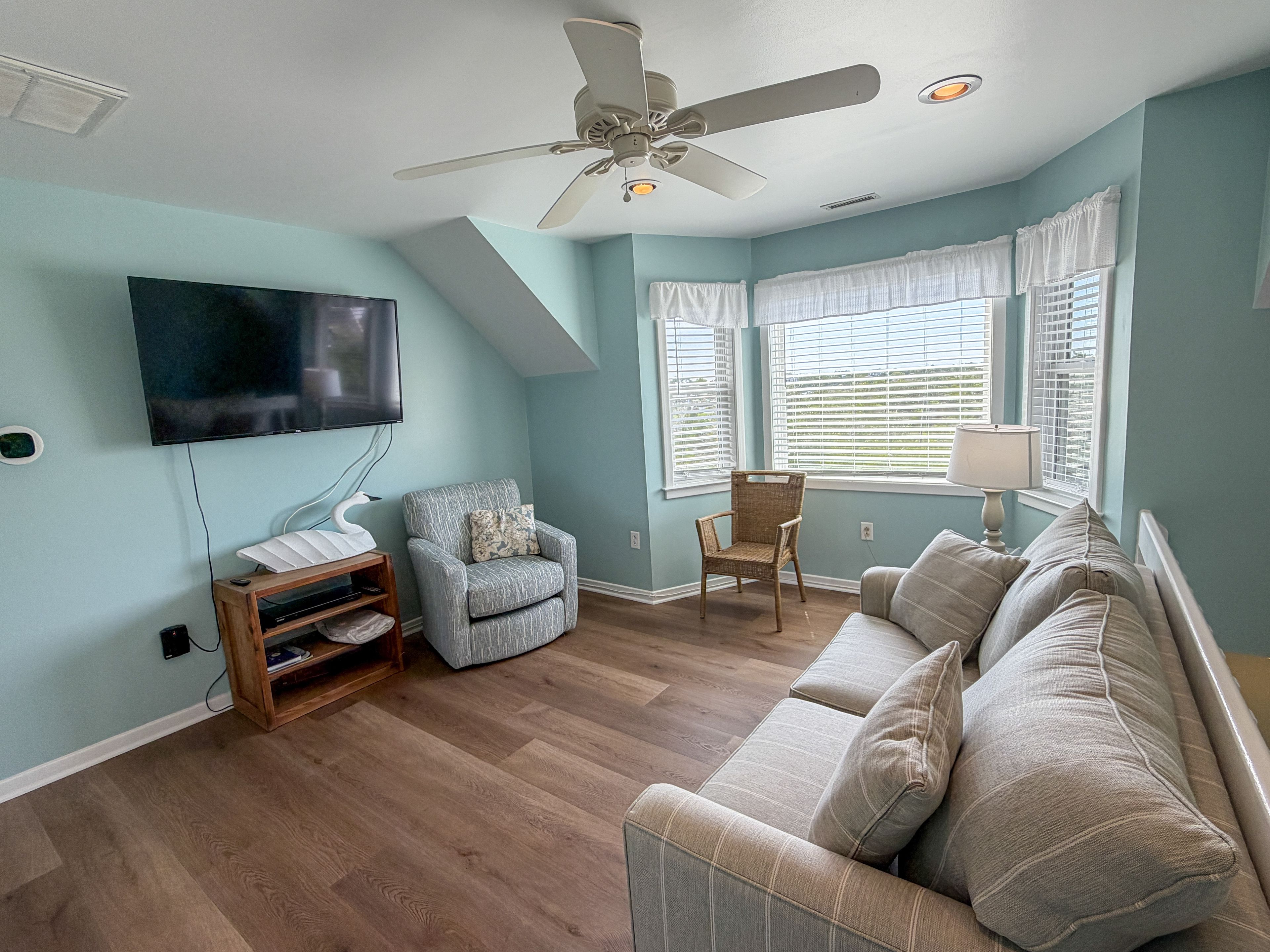 Ein Wohnzimmer mit Deckenventilator, einem Flachbildfernseher, einer hellblauen Wand, Holzboden, einem grauen Sofa und einem Korbstuhl.