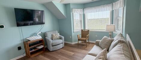Ein Wohnzimmer mit Deckenventilator, einem Flachbildfernseher, einer hellblauen Wand, Holzboden, einem grauen Sofa und einem Korbstuhl.