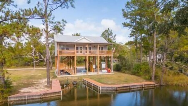 A two-story house with a dock extending into a body of water, surrounded by trees.