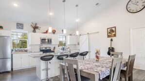 A modern kitchen with white cabinets, a central island with bar stools, a dining table with chairs, and a guitar in the background.