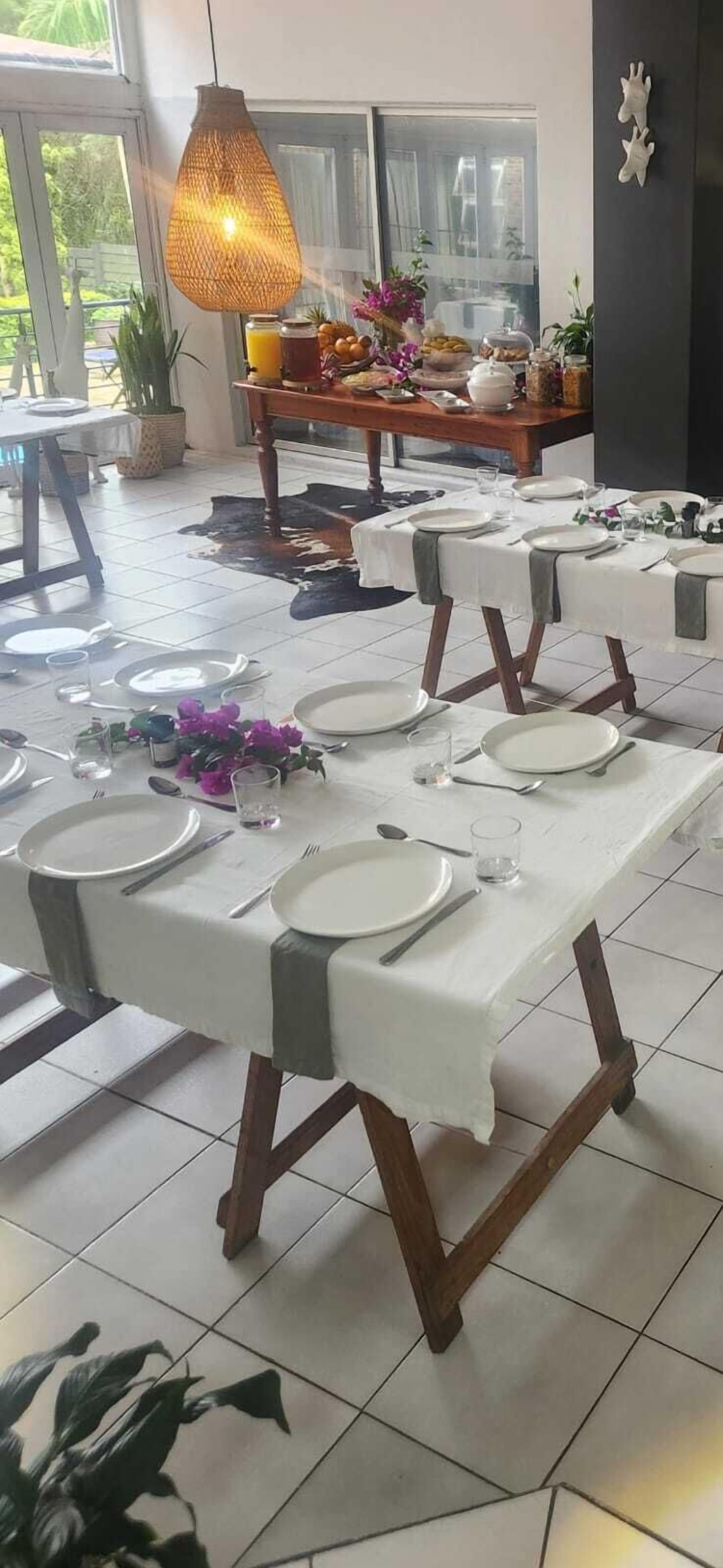 A dining area with multiple tables set for guests, featuring white tablecloths and wooden chairs.