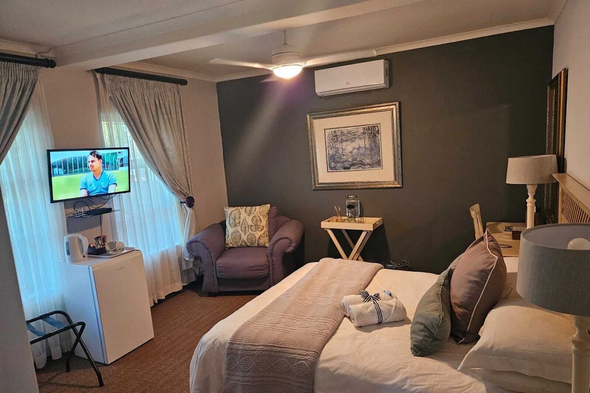 A hotel room with a bed, a desk with a TV, a chair, a small table with a lamp, and a framed picture on the wall.