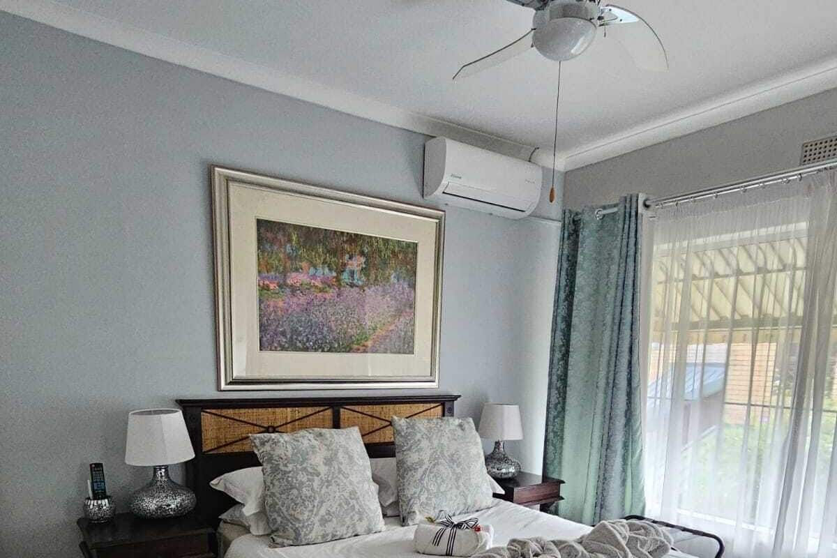 A neatly made bed with a white comforter, a decorative pillow, and a blanket. There is a framed painting above the bed, a ceiling fan, and a window with curtains.