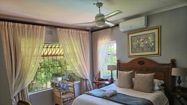 A hotel room with a bed, a desk, a chair, a ceiling fan, and an air conditioning unit. - Hillton Manor Guest House (Richards Bay)