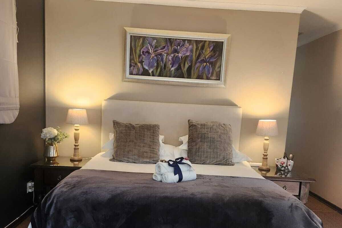 A neatly made bed with a dark blanket, two pillows, and a folded towel on a headboard. There is a ceiling fan above and a painting of flowers on the wall.