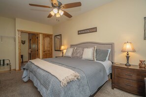 A bedroom with a bed, two bedside lamps, a ceiling fan, a wooden door, and a wall sign. - Creekside Bliss | 2BR Cabin • Walk to Downtown • Patio + Fireplace + WIFI (Gatlinburg)