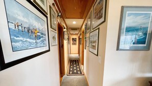 A hallway with framed pictures on the walls, a carpeted floor, and wooden paneling on the ceiling. - Bluefin: Nautical and eclectic artwork, and amazing marsh views. (Ocracoke)