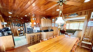 A wooden interior with a dining table, kitchen area, and a ceiling fan. - Bluefin: Nautical and eclectic artwork, and amazing marsh views. (Ocracoke)