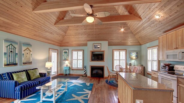 A spacious living area with a high ceiling, wooden beams, a fireplace, a dining table, and a kitchen with a microwave. - Cabana III:  Perfect getaway for two that offers a shared pool. (Ocracoke)