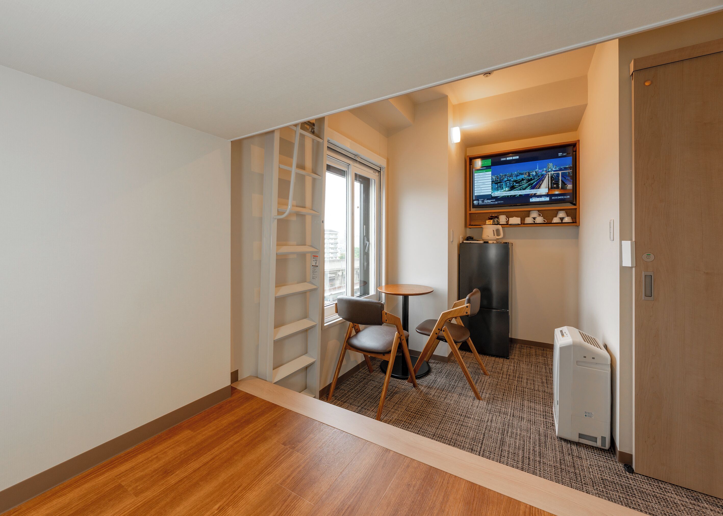 A hotel room with a wooden floor, a small round table with two chairs, a TV mounted on the wall, and a refrigerator.