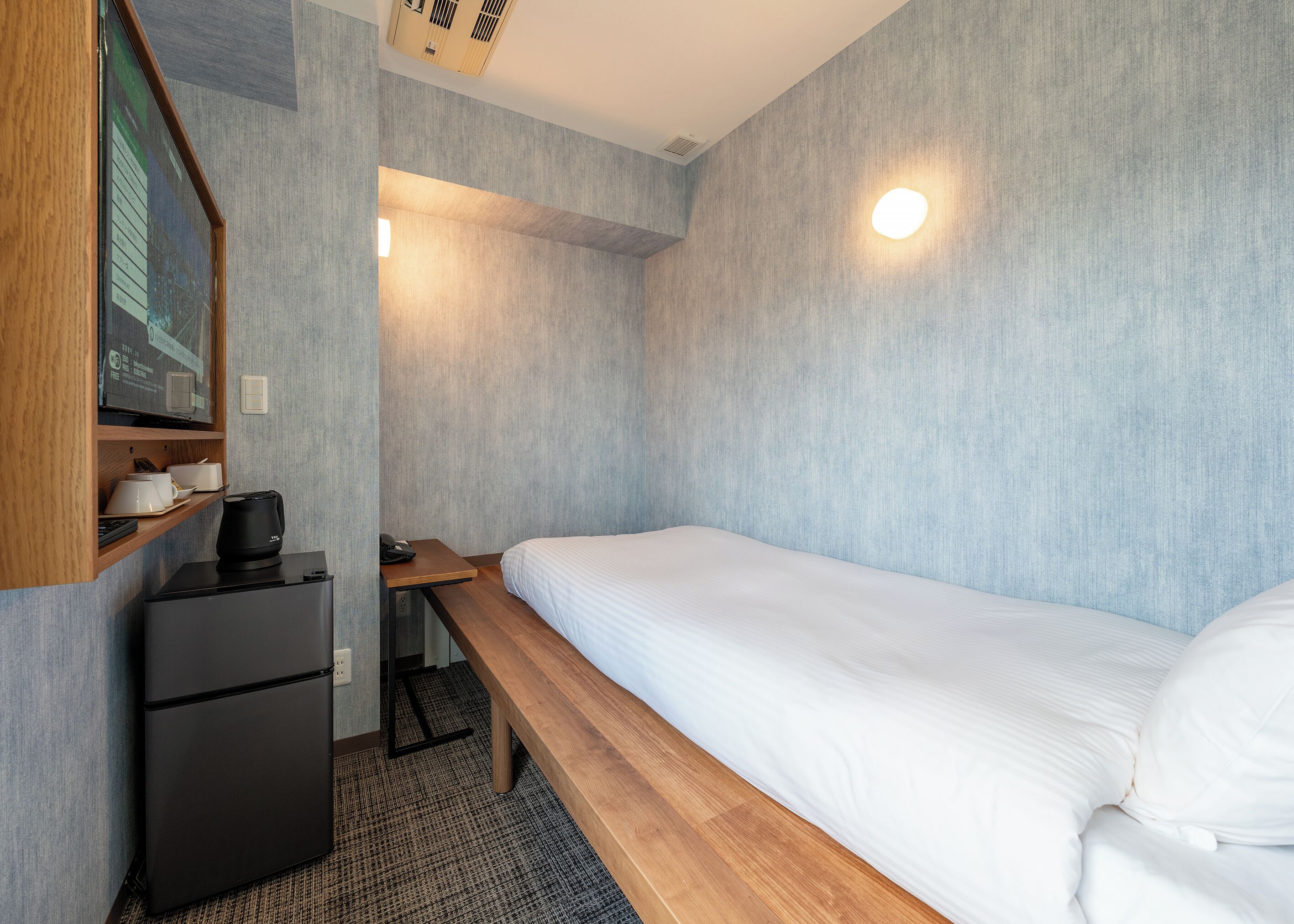 A compact hotel room with a single bed, a small desk with a computer and a coffee maker, and a built-in refrigerator.