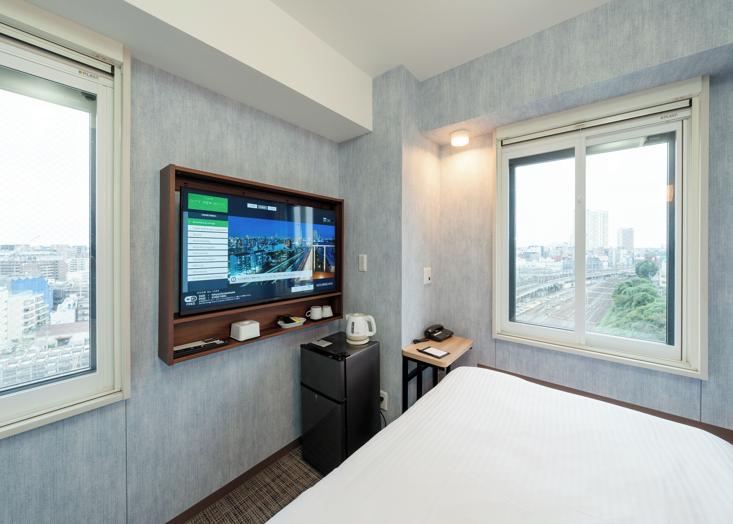 A hotel room with a bed, a TV mounted on the wall, a small desk with a phone and a cup, and a window with a city view.