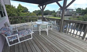 A wooden deck with a round table and chairs, a cushioned bench, and a railing overlooking a body of water.