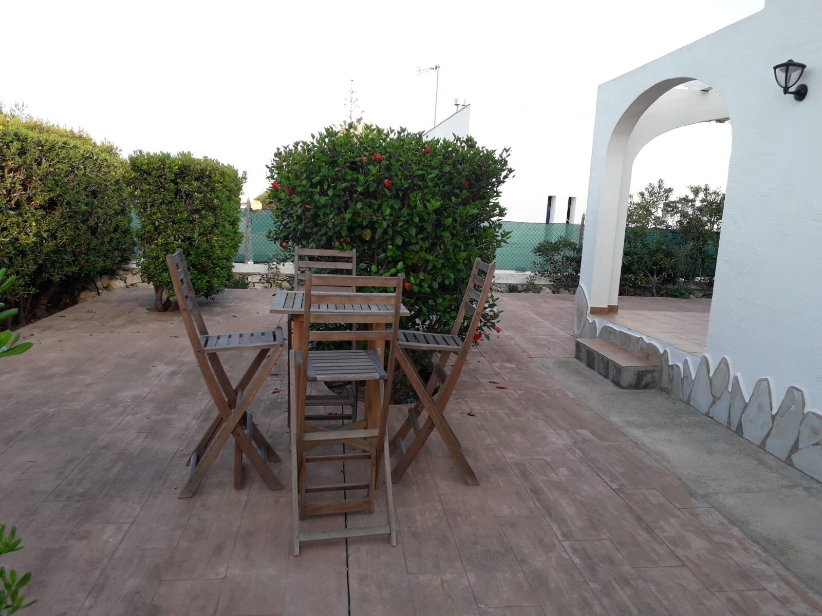 A patio with a wooden table and chairs, surrounded by greenery and a white wall with an archway.
