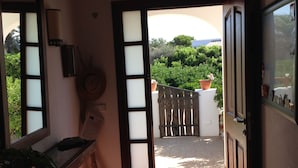 Un couloir carrelé mène à une porte ouverte. Un pot de fleurs se trouve sur la gauche et une barrière en bois est placée à l’extérieur.