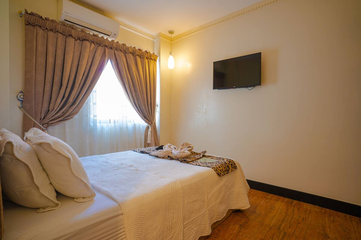 A neatly made bed with a white comforter and pillows, a flat-screen TV mounted on the wall, and a window with curtains.