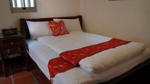 A neatly made bed with a white comforter and a red blanket, a wooden nightstand with a lamp, and a window with blinds. - Teapot House (Jinning)