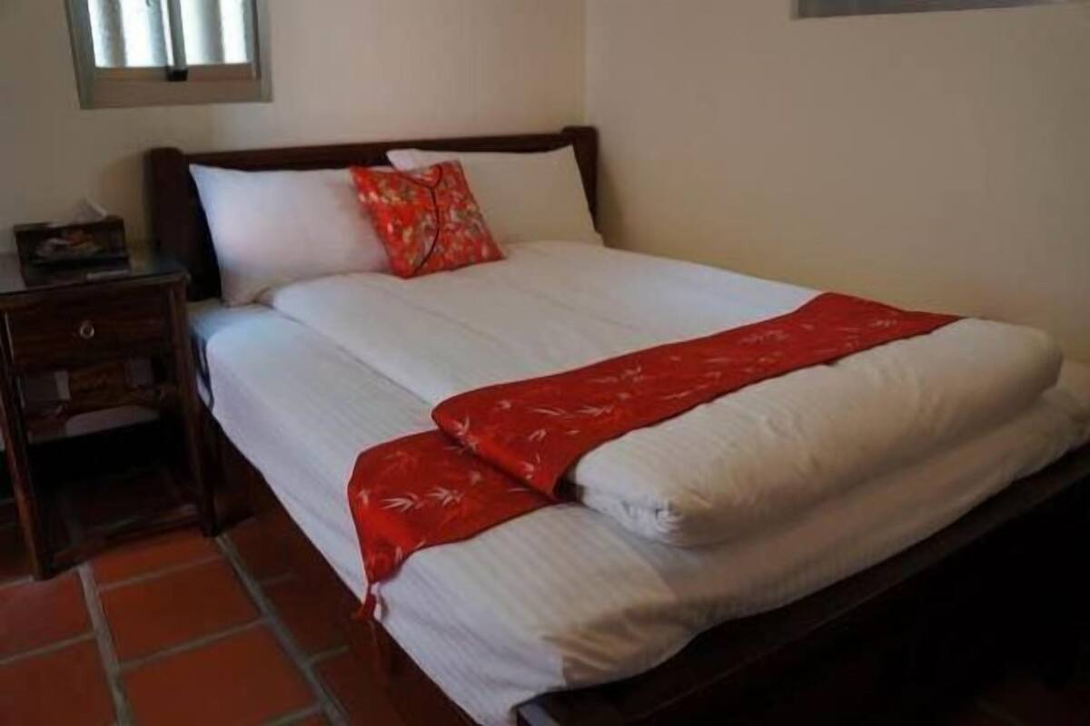 Una cama bien hecha con edredón blanco y manta roja, una mesita de noche de madera con lámpara, y una ventana con persianas.
