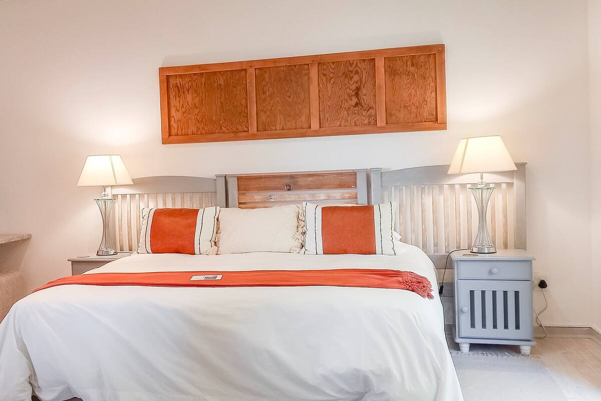 Un dormitorio con una cama, dos mesitas de noche con lámparas, una silla, un espejo redondo y un panel de madera sobre la cama.