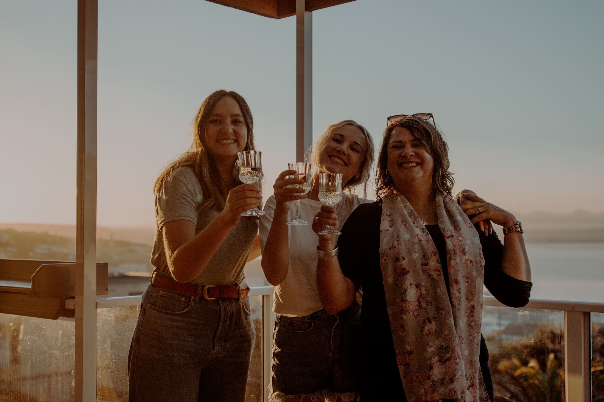 Three people toasting with glasses, standing in a room with a view of a body of water and a clear sky.