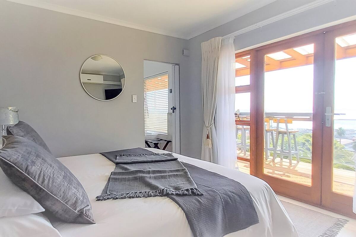 Un dormitorio con cama, lámpara de noche, espejo y vistas al exterior a través de un ventanal corredero.