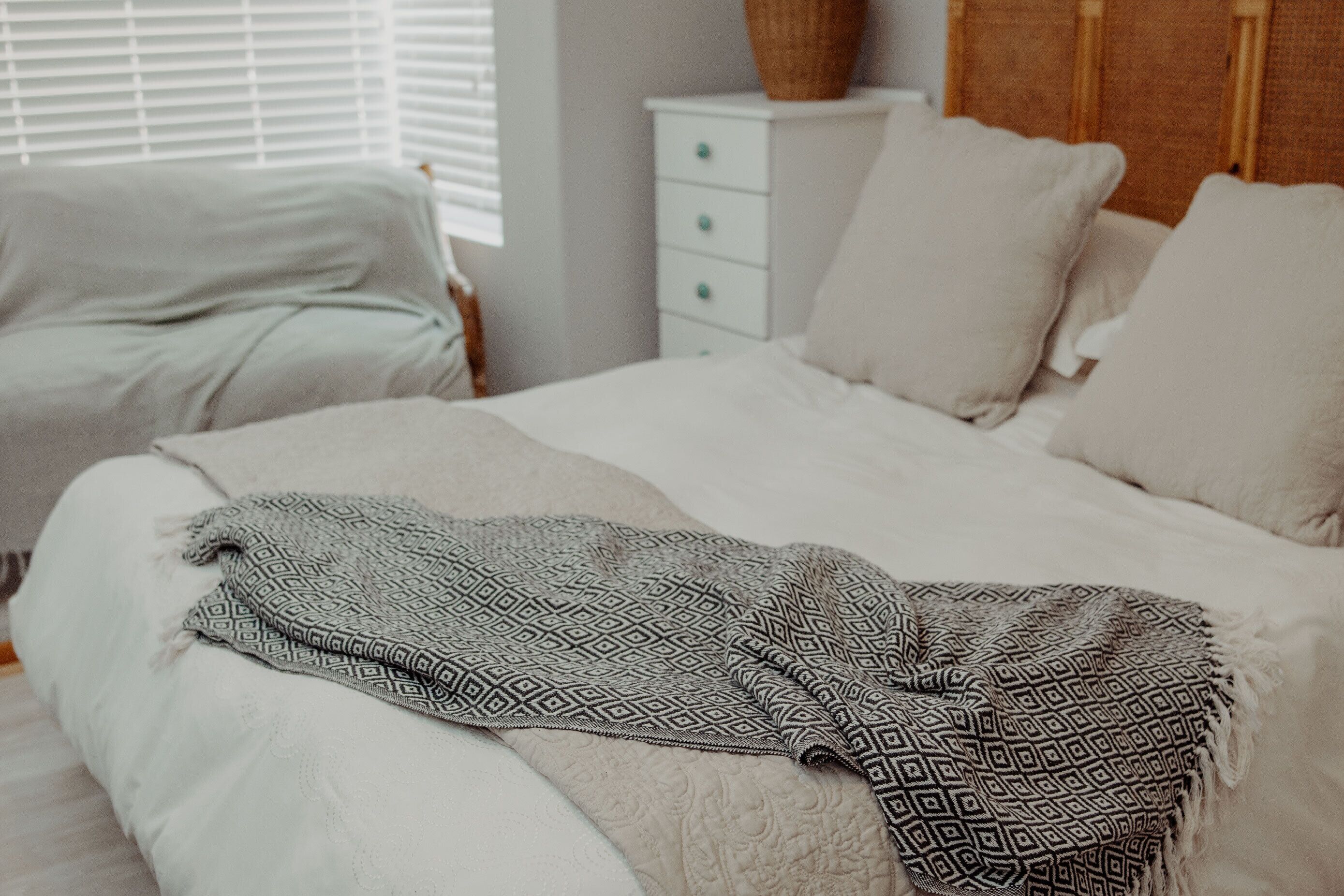 A neatly made bed with a patterned throw blanket, a white bedside table with drawers, and a wicker basket on top.