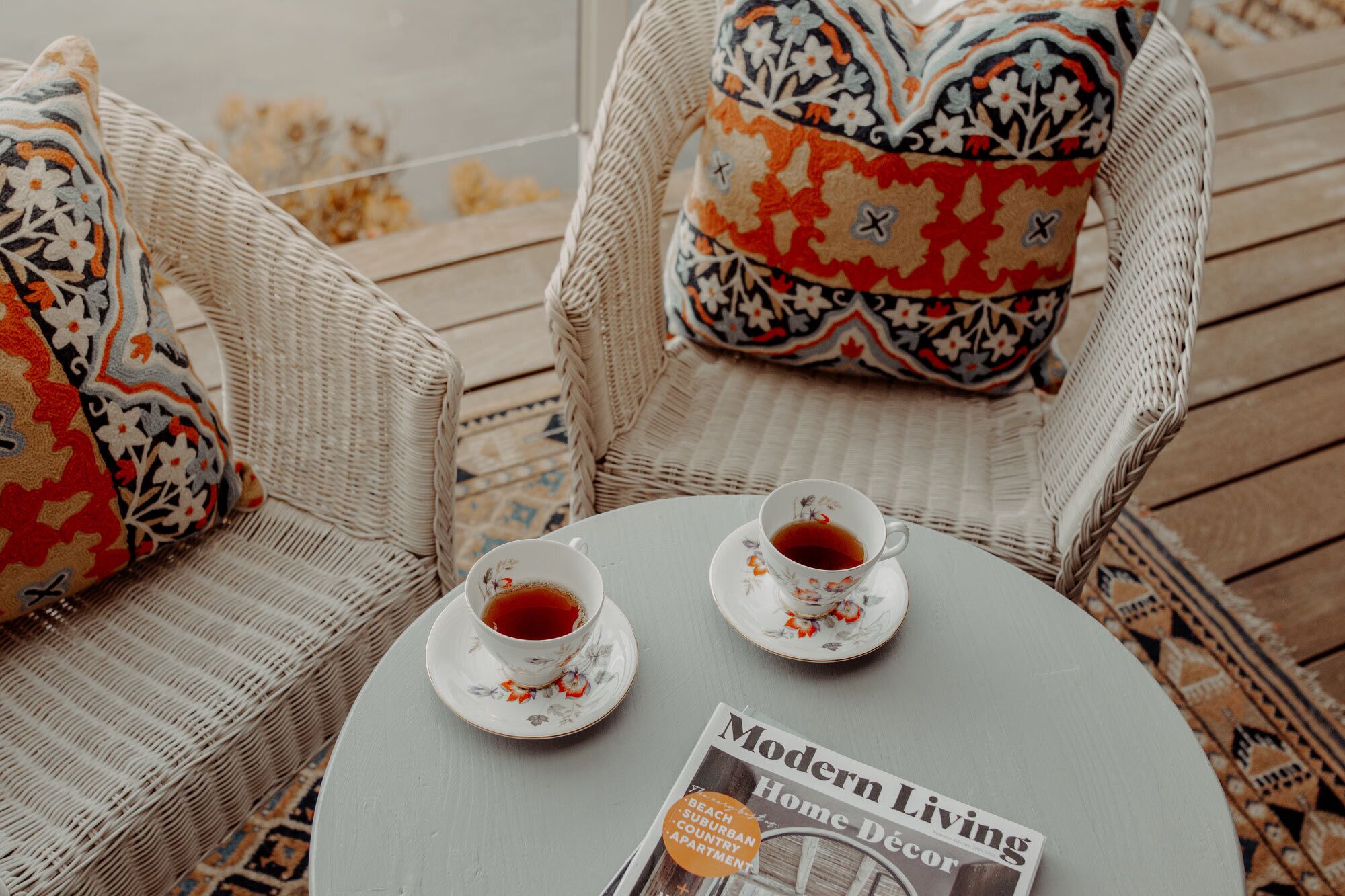 Two wicker chairs with patterned cushions, a round table with two teacups and saucers, and a magazine titled "Modern Living Home Decor".