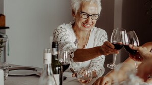 An elderly woman at a table with wine glasses, a bottle of wine, and a glass of water.