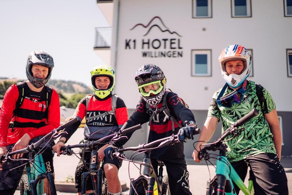 Four cyclists in front of a building with a sign that reads 'K1 Hotel Willingen.'