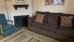 A living room with a brown sofa, teal armchair, patterned rug, fireplace, framed artwork, and a tall floor lamp. - Olde Gatlinburg 203 is a beautiful condo with private balcony located in downtow (Gatlinburg)