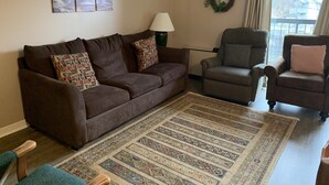 A living room with a brown sofa, patterned cushions, a patterned rug, a green armchair, and a view through a window. - Olde Gatlinburg 203 is a beautiful condo with private balcony located in downtow (Gatlinburg)
